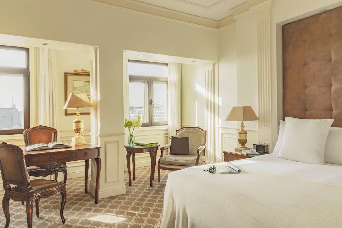 Une chambre d’hôtel avec un grand lit, un bureau avec une chaise, une petite table avec une lampe et une fenêtre avec des rideaux.