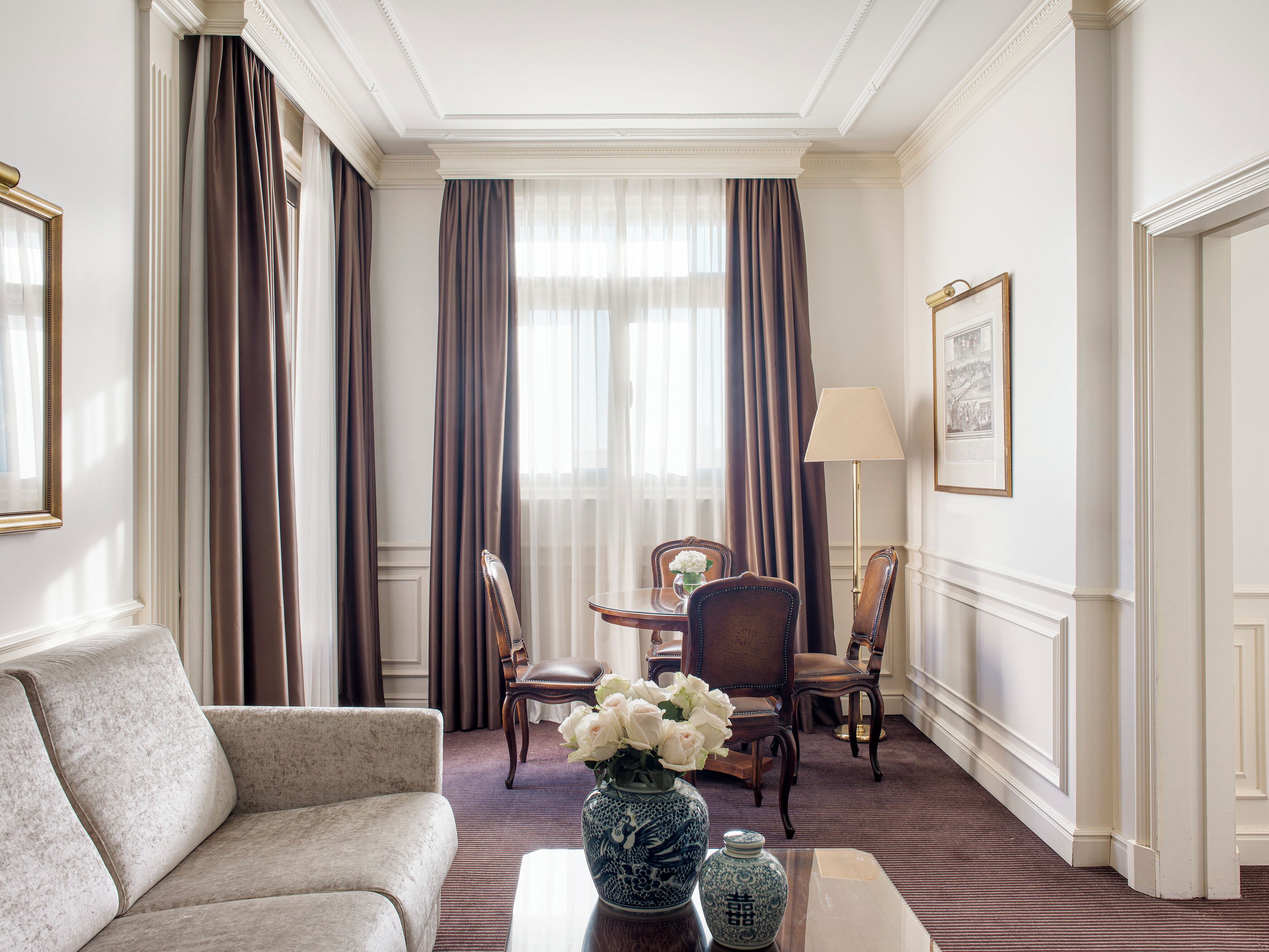Una habitación de hotel con sofá, una mesa de comedor con sillas, un jarrón con flores y una lámpara.