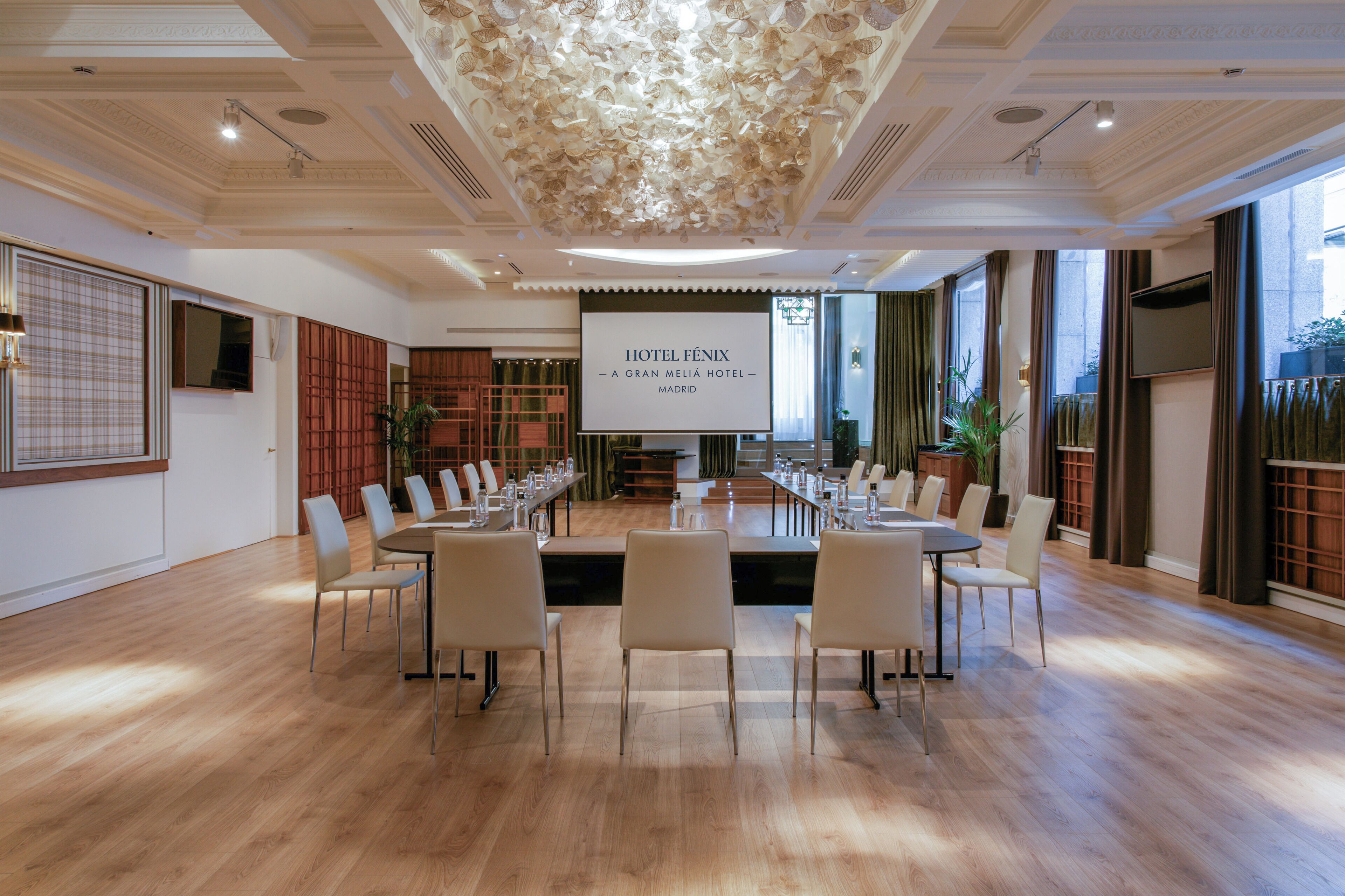 Una grande sala conferenze con un lungo tavolo e sedie, uno schermo che mostra 'HOTEL FÉNIX - A GRAN MÉLIA HOTEL - MADRID' e ampie finestre con tende.