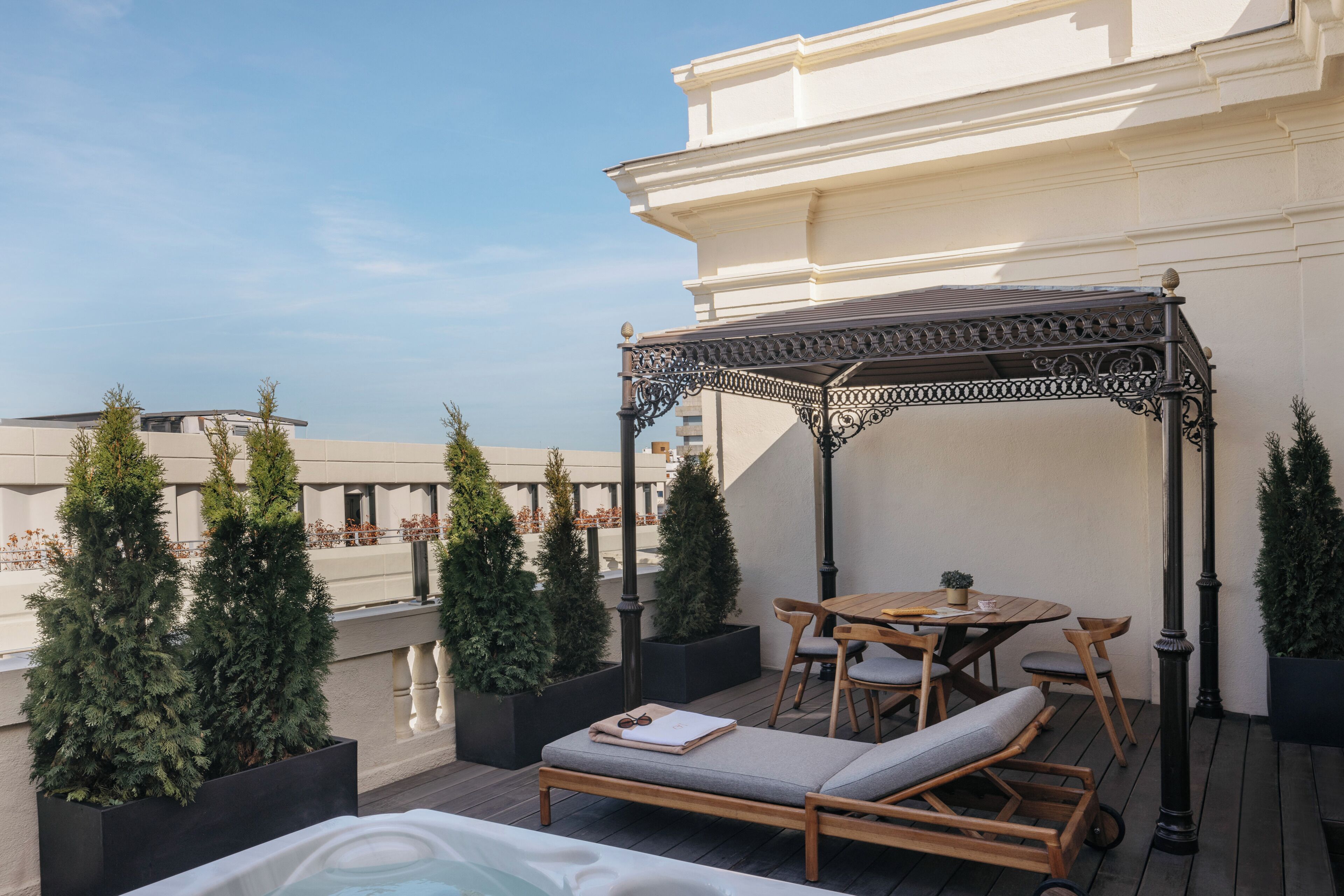 Una terraza en la azotea con una silla de descanso de madera, una mesa con sillas, una pérgola y árboles de hoja perenne en macetas.