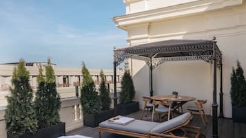 Una terraza en la azotea con una silla de descanso de madera, una mesa con sillas, una pérgola y árboles de hoja perenne en macetas.