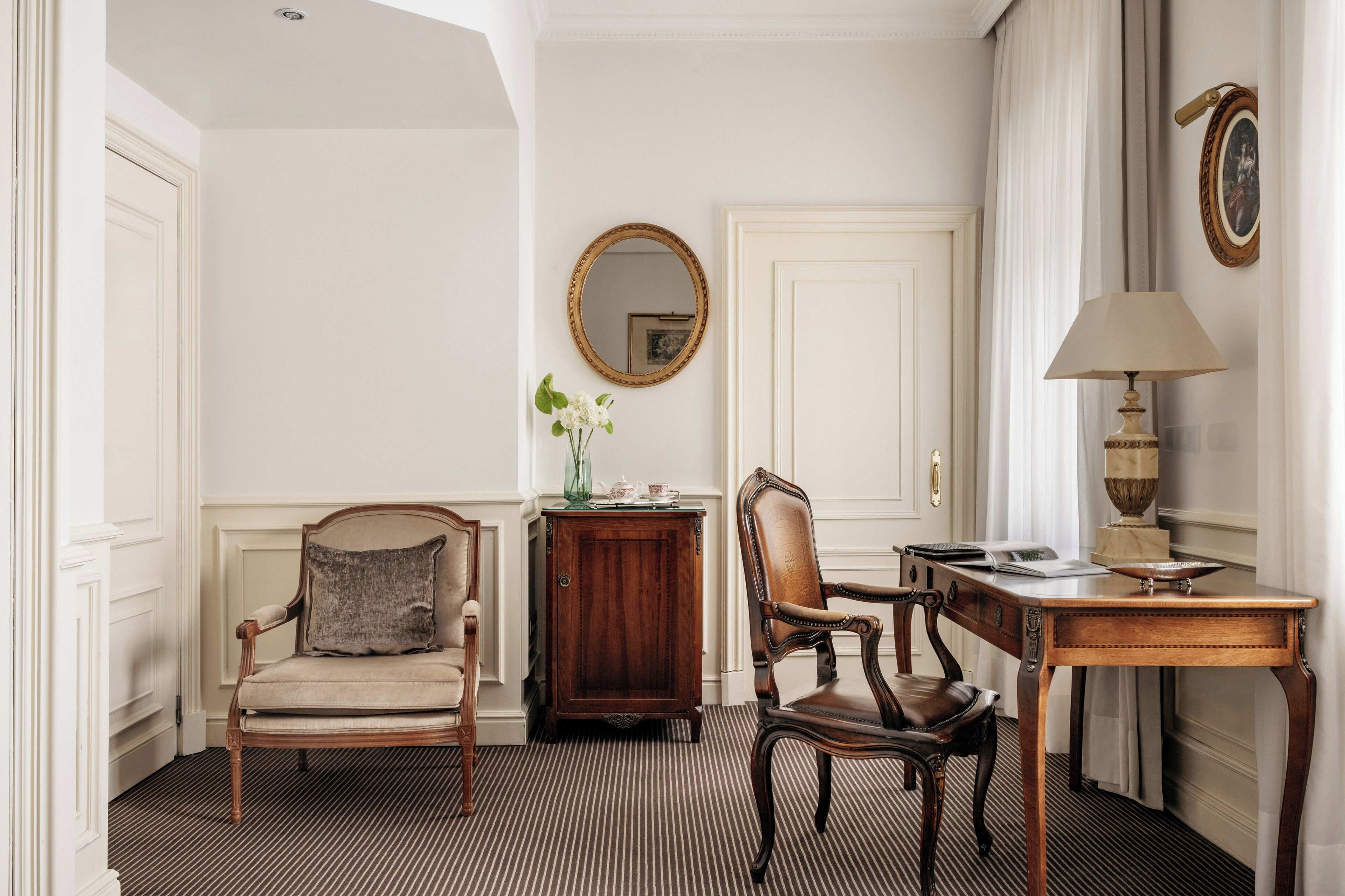 A room with a striped carpet, a wooden cabinet, a round mirror, a table with a lamp, and two chairs.