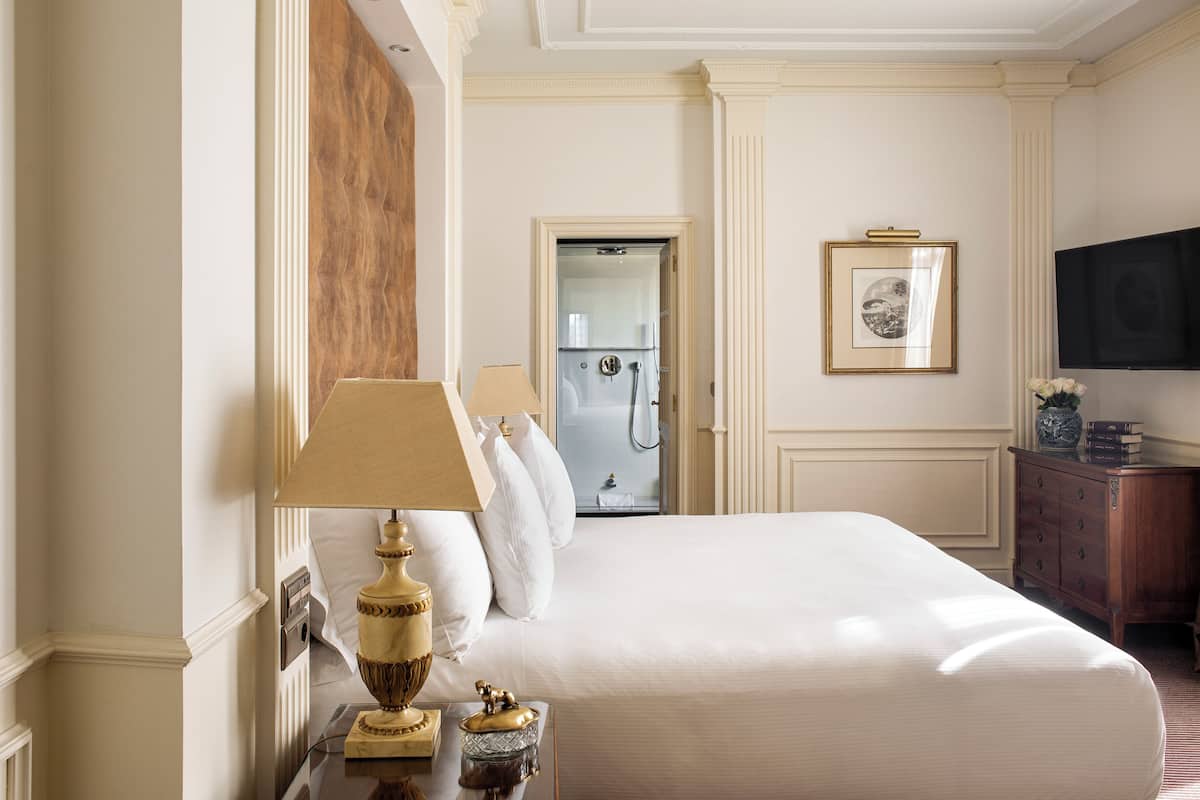 Une chambre d’hôtel avec un grand lit, une tête de lit en bois, une table de chevet avec une lampe, une commode, un téléviseur et une salle de bain avec douche.