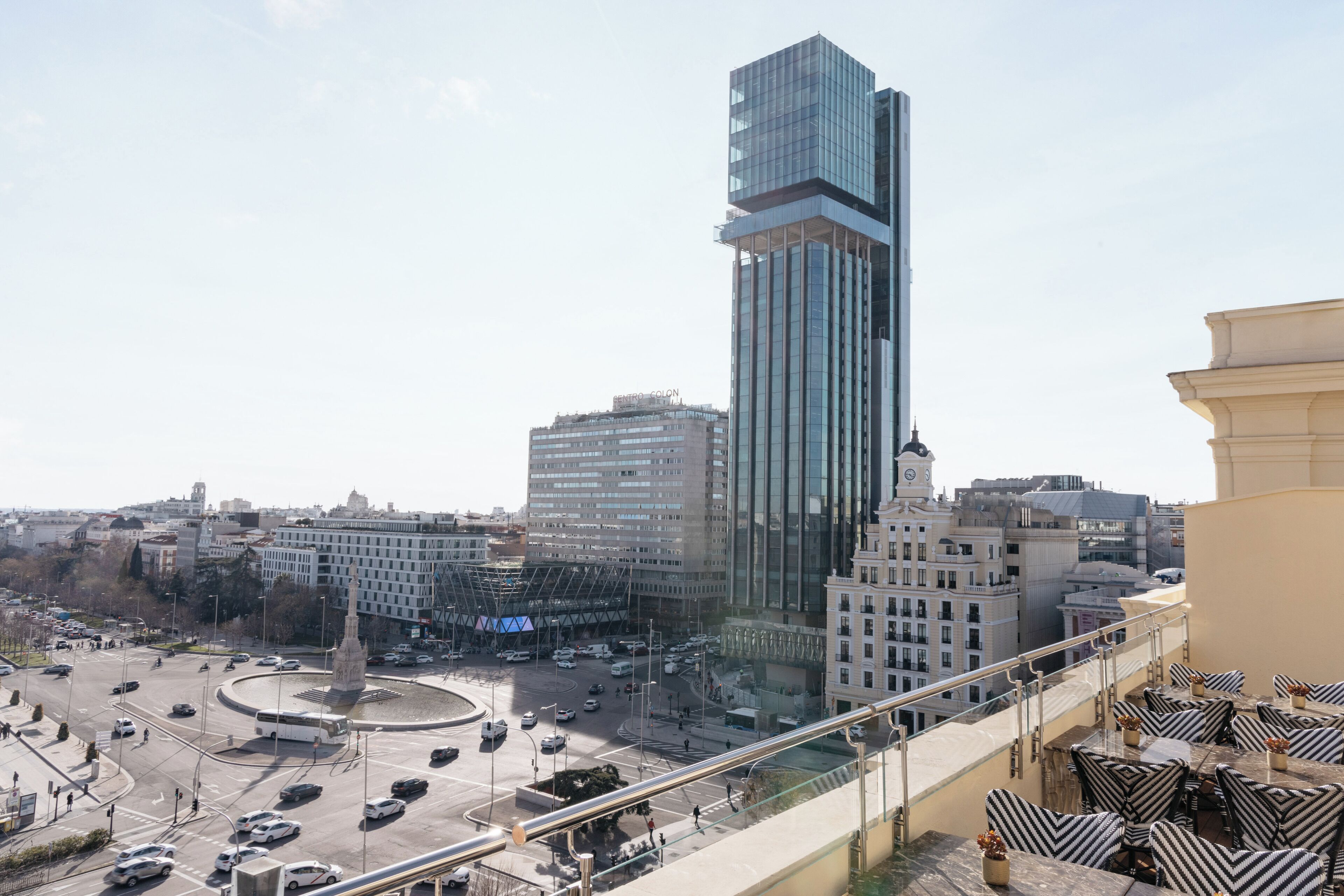 Um edifício alto com fachada de vidro moderna, uma rotatória com trânsito e um café com mesas ao ar livre.