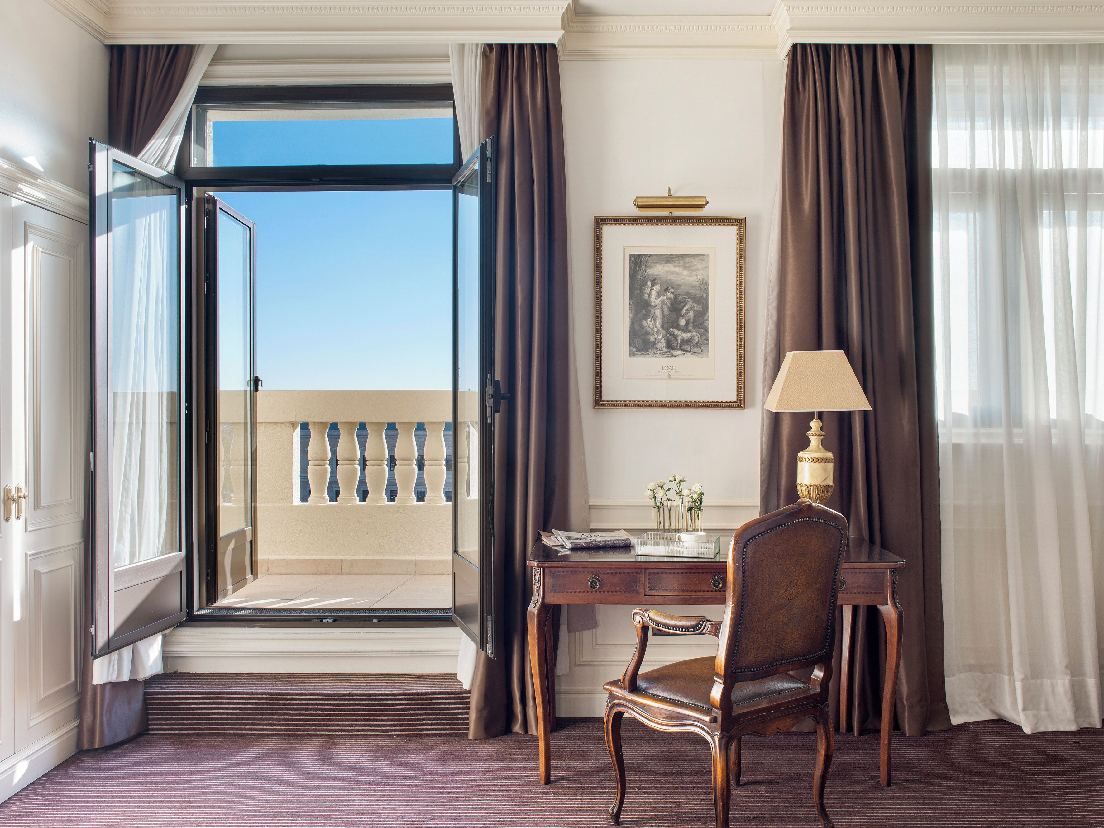 A hotel room with a desk, chair, and a view of the balcony.