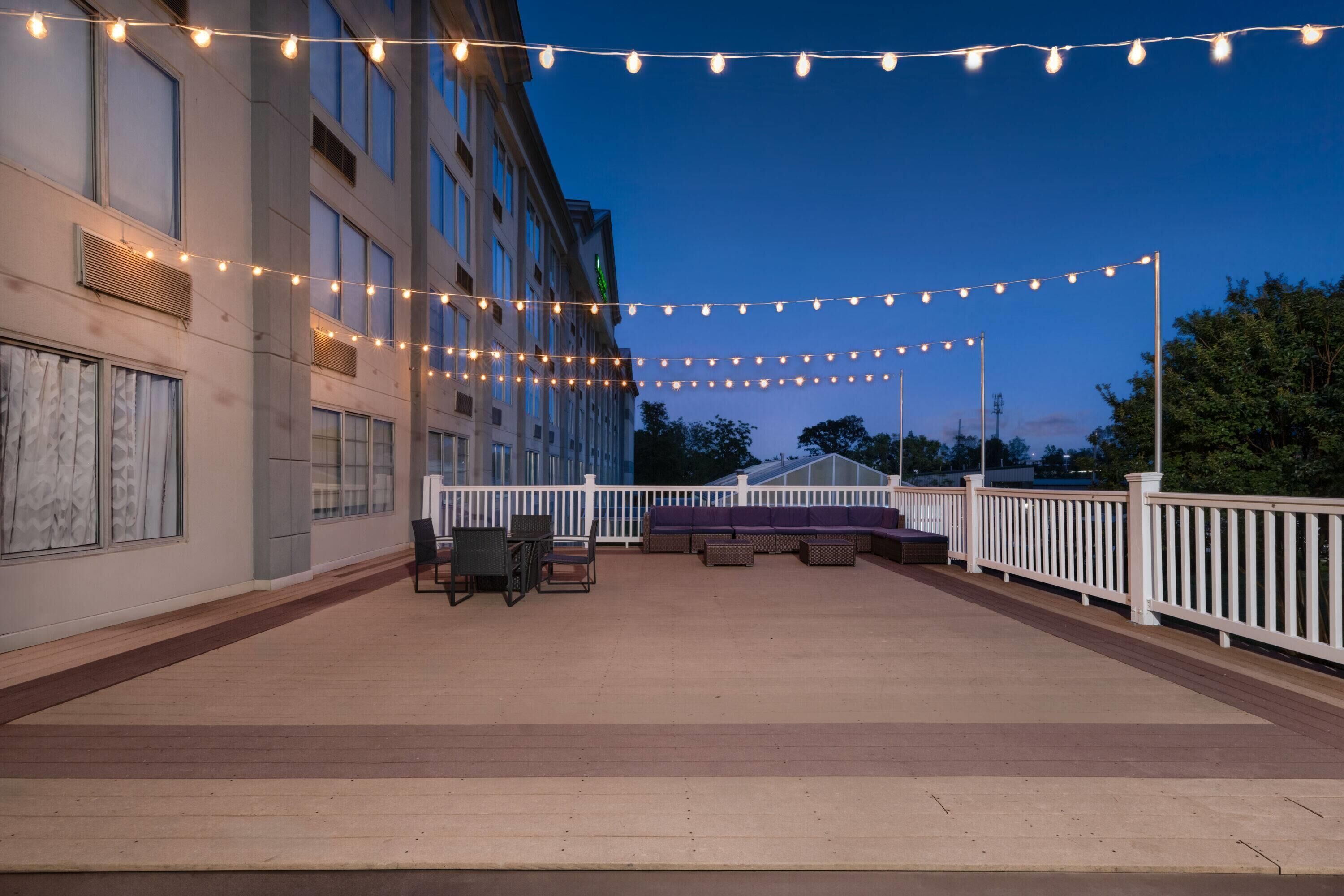 Un patio al aire libre con luces colgantes, mobiliario exterior y barandal blanco.