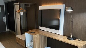 Habitación de hotel moderna con una televisión de pantalla plana grande montada en un mueble de madera, un escritorio con lámpara, un armario y un cesto de basura.