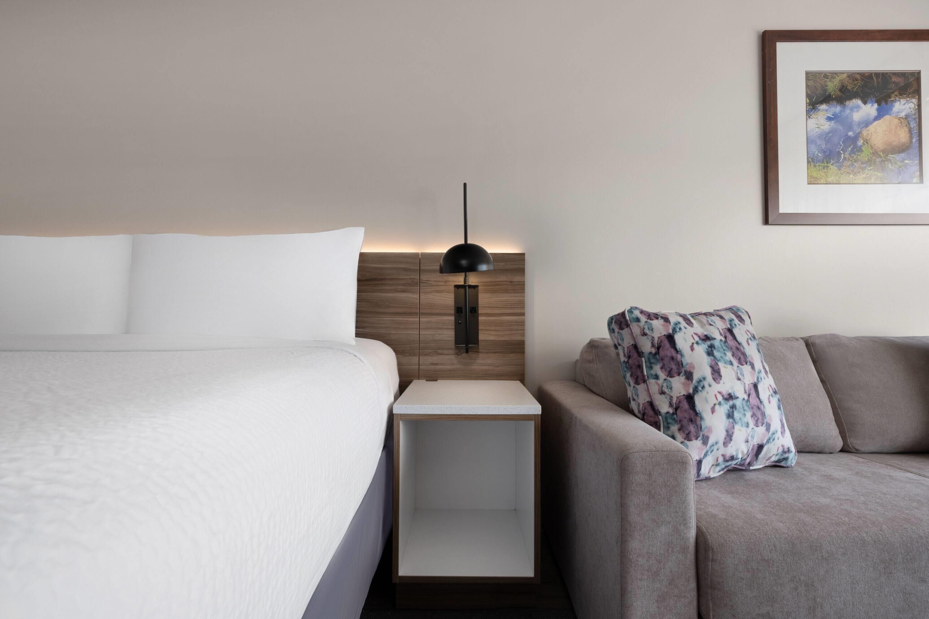A hotel room with a bed, a nightstand, a lamp, a sofa, and a framed picture on the wall.