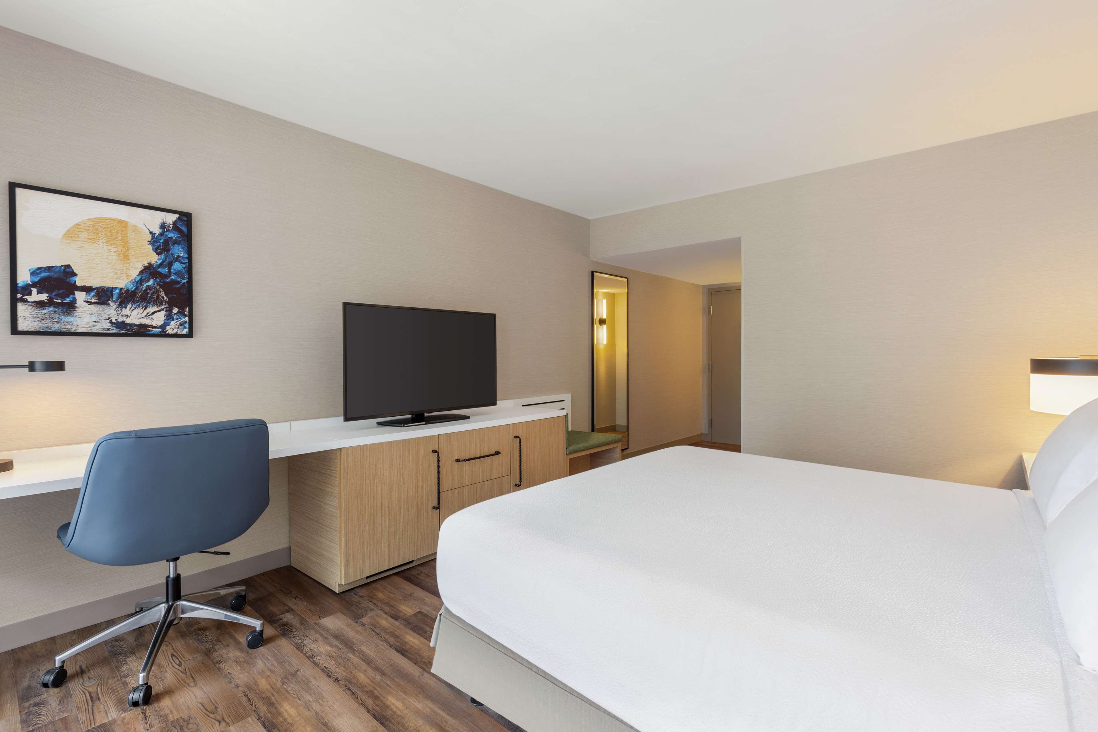 A hotel room with a bed, a desk with a chair, a television, and a painting on the wall.