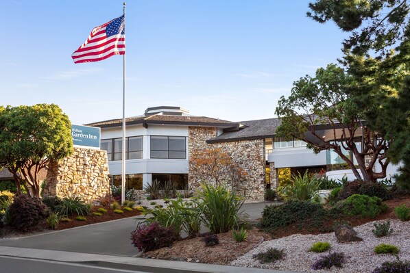 A modern hotel with a stone facade, a flagpole with an American flag, and a landscaped garden. - Hilton Garden Inn Monterey (Monterey)