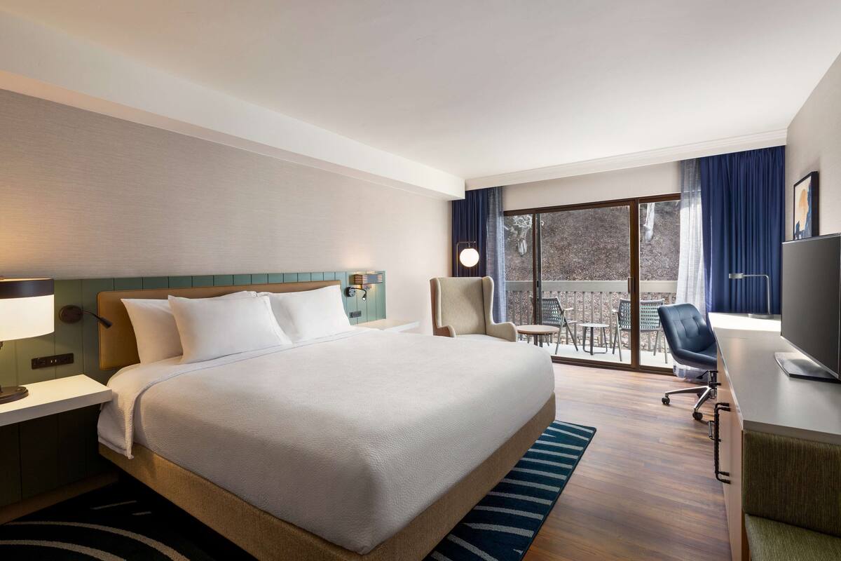 A hotel room with a large bed, a desk with a computer, a chair, and a view of the outdoors through a sliding glass door.