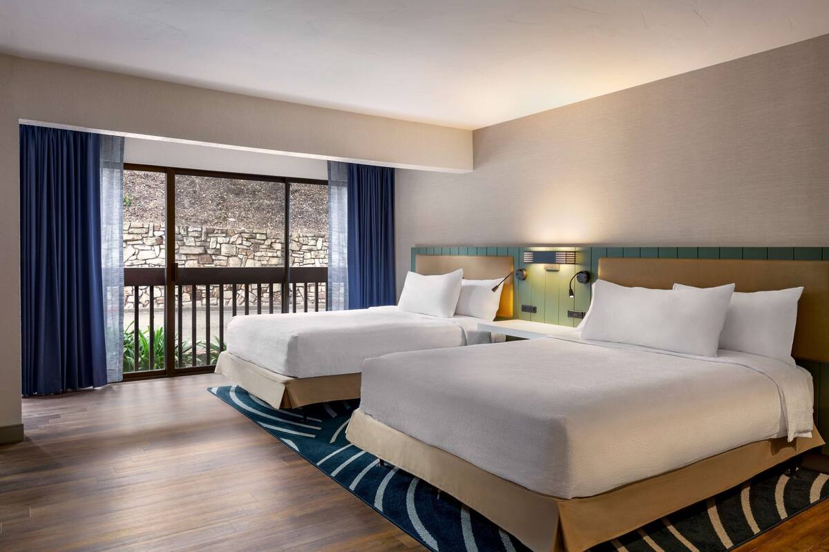 A hotel room with two beds, a balcony with a stone wall, and a view of greenery.