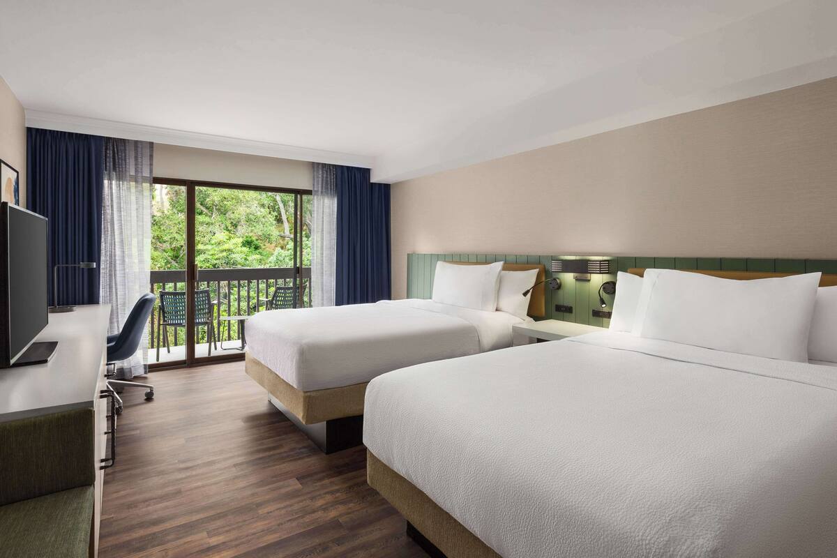 A hotel room with two beds, a desk, a chair, and a TV. There is a balcony with a view of greenery.