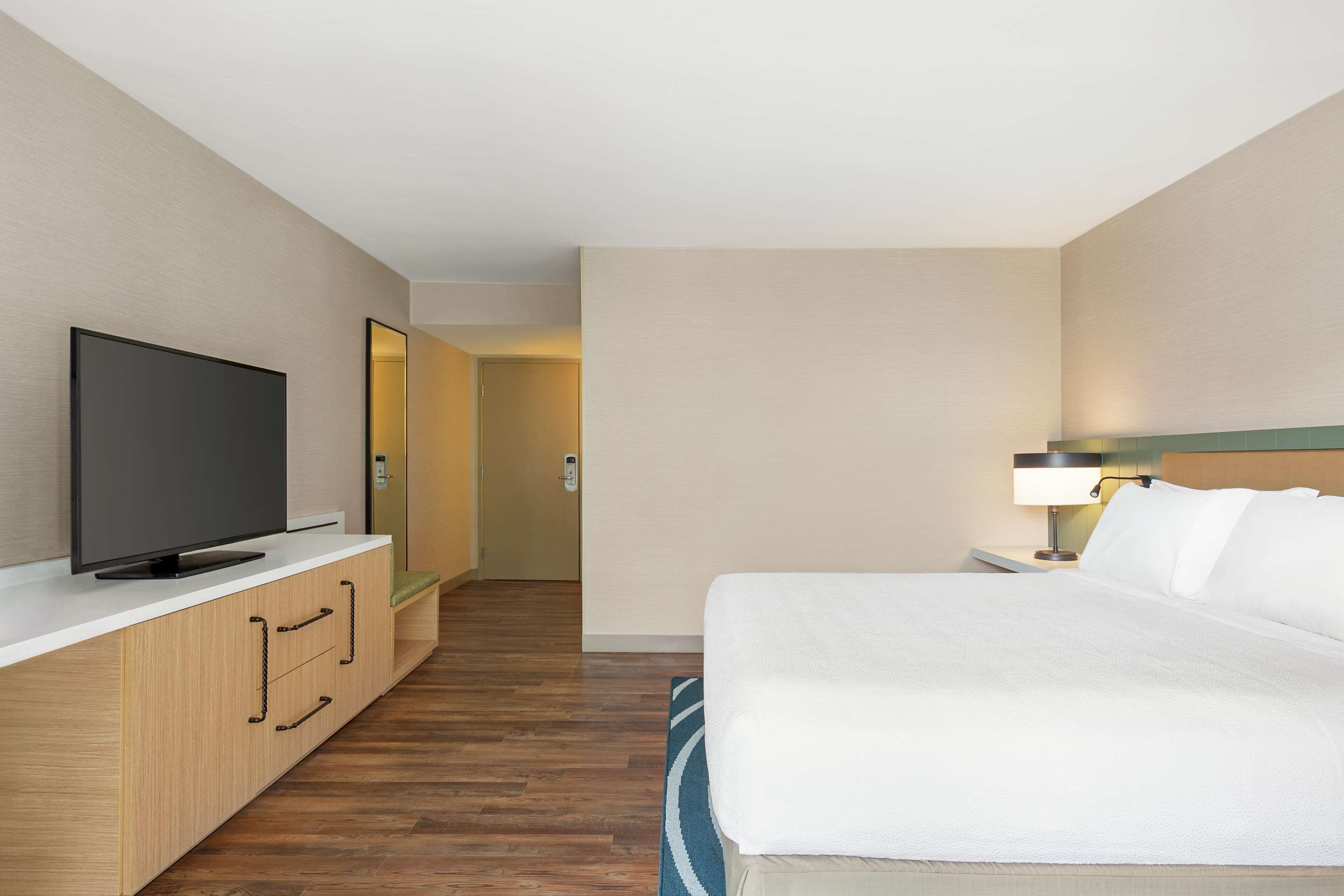 A hotel room with a bed, a TV on a wooden cabinet, a nightstand with a lamp, and a mirror on the wall.