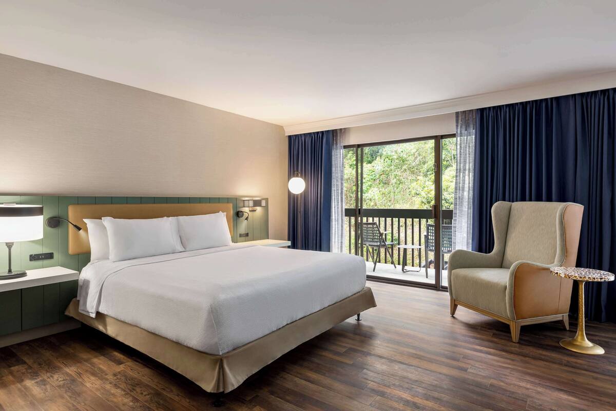 A hotel room with a large bed, a chair, a small table, a nightstand, and a view of the outdoors through a balcony.