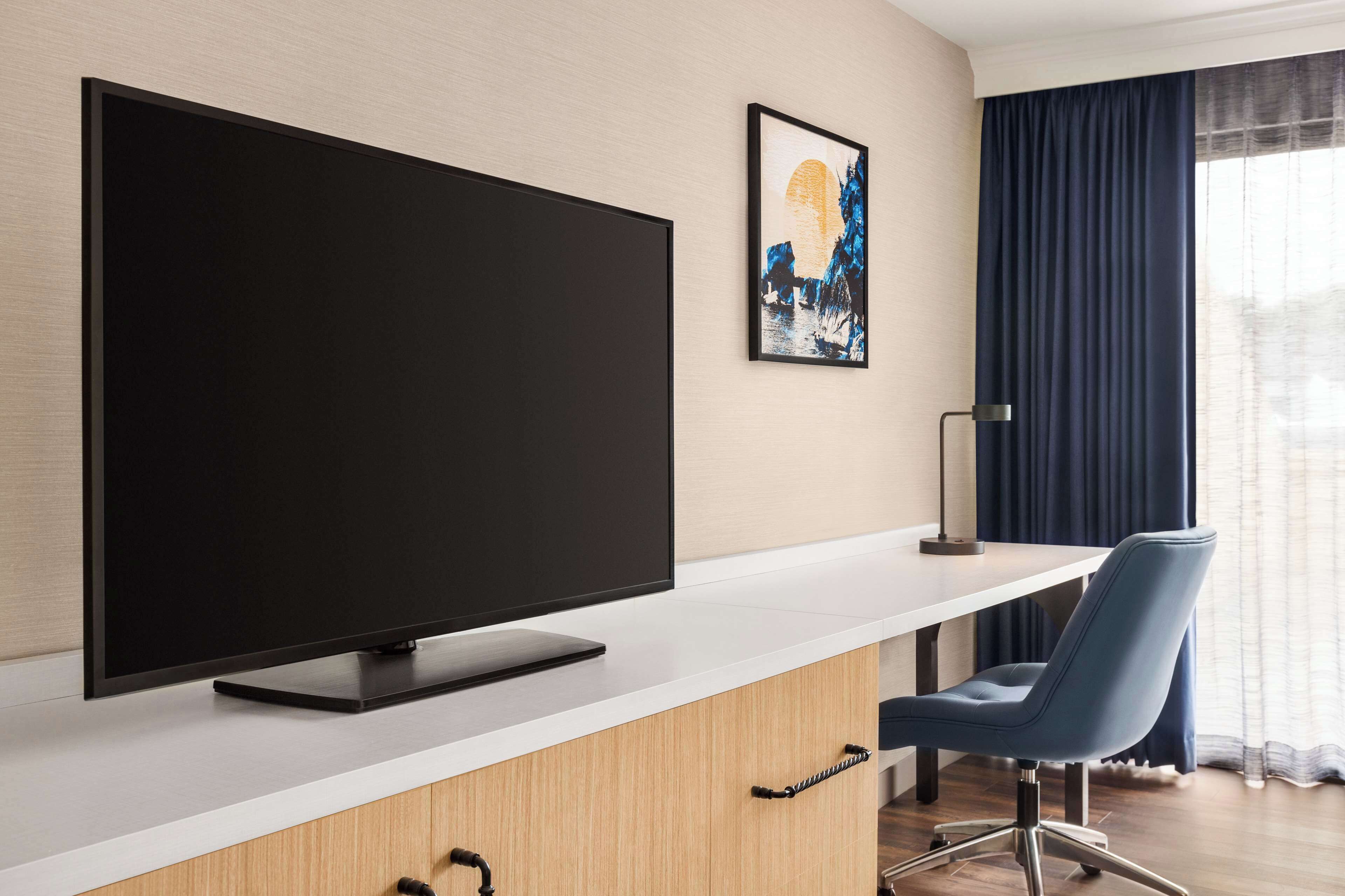 A modern hotel room with a large flat-screen TV on a white console, a blue chair, and a painting on the wall.