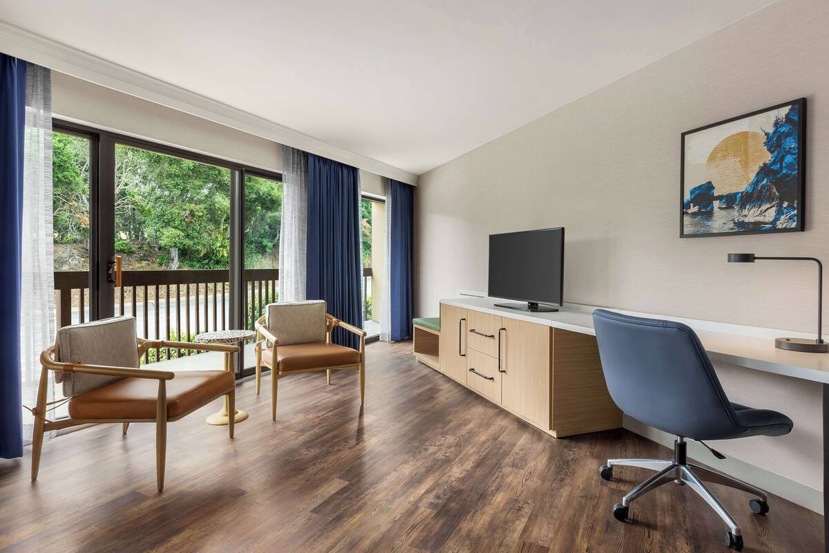A modern room with a wooden floor, a desk with a computer, two chairs, a TV, and a large window with curtains.