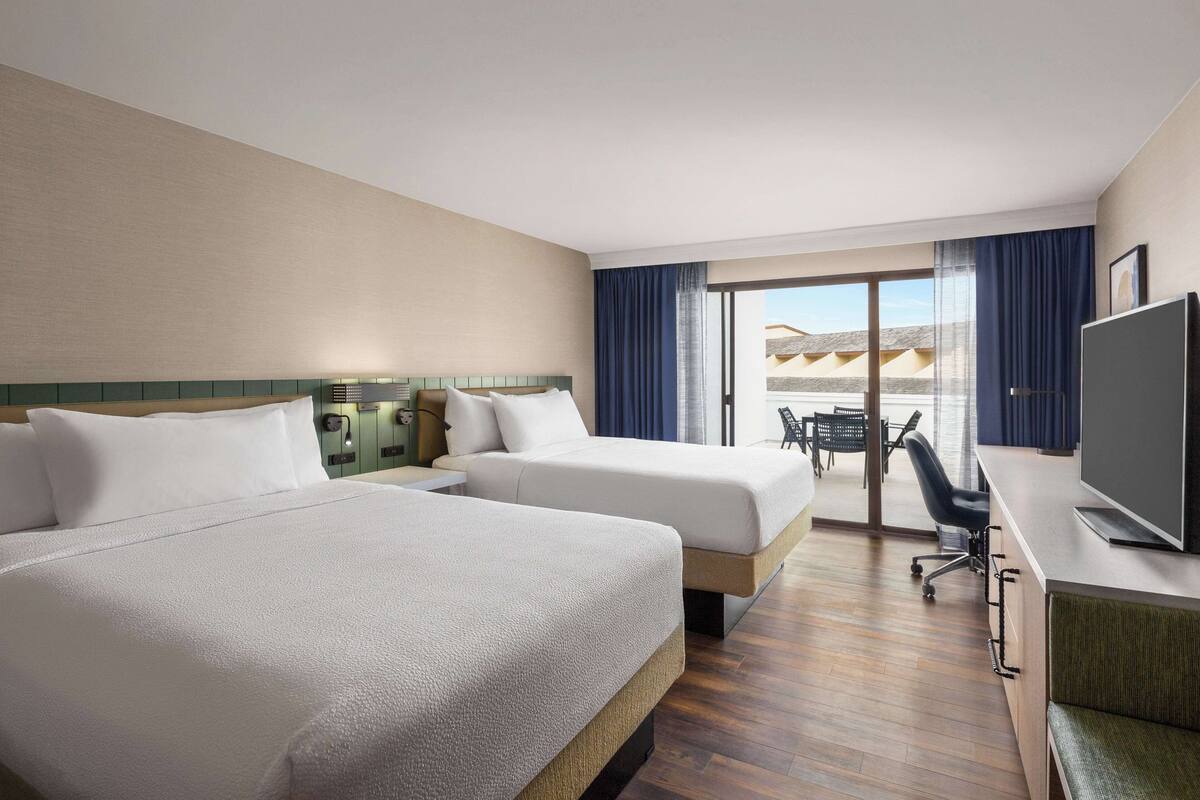A hotel room with two beds, a TV, a desk, and a balcony with a view.