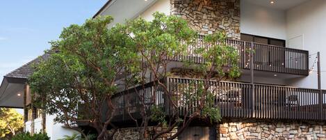 A modern two-story house with a stone facade, a balcony with metal railings, and lush greenery.