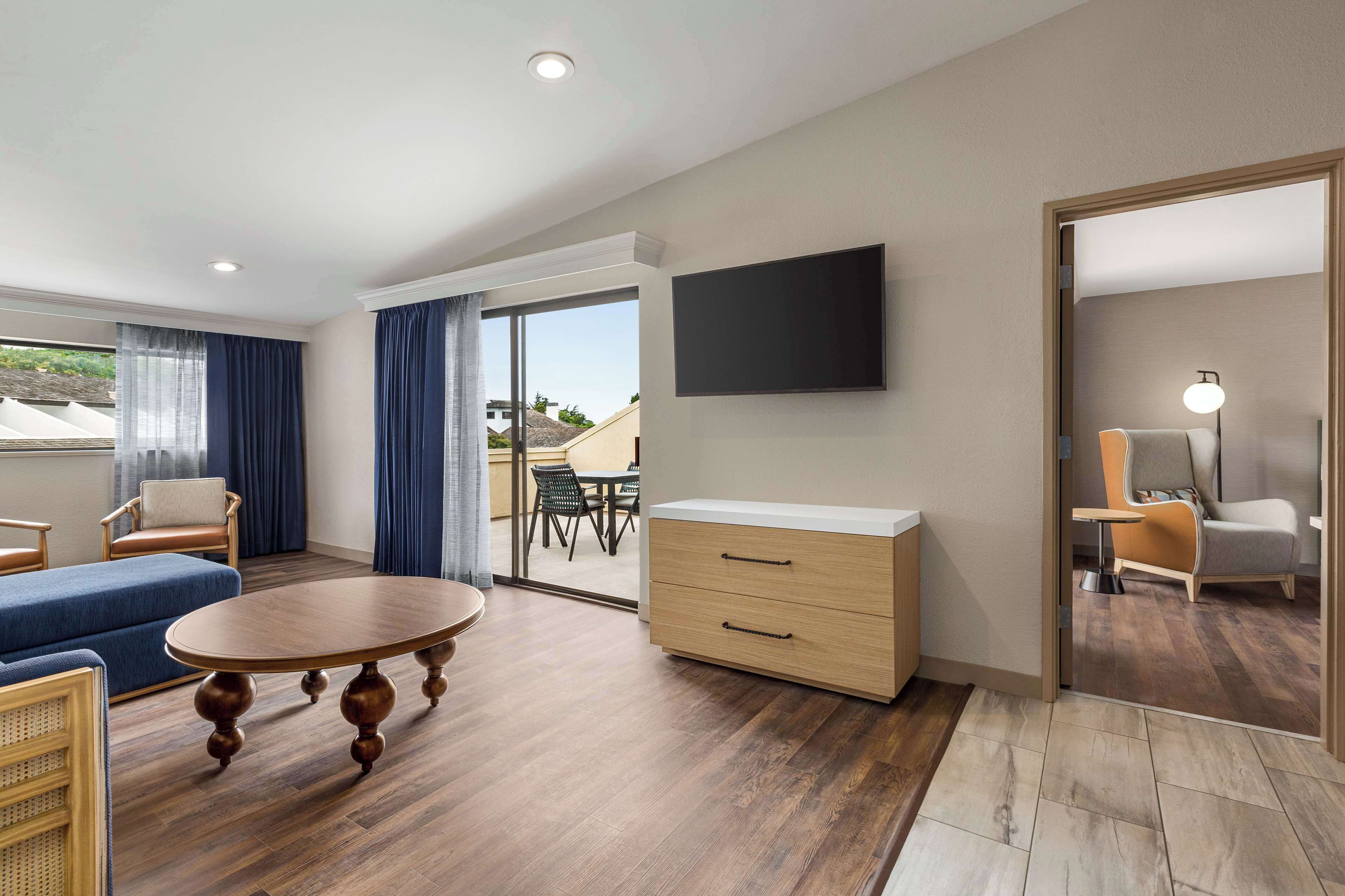 A modern hotel room with a wooden floor, a round table, a sofa, a TV, and a sliding glass door leading to a balcony.