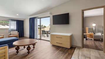 A modern hotel room with a wooden floor, a round table, a sofa, a TV, and a sliding glass door leading to a balcony.
