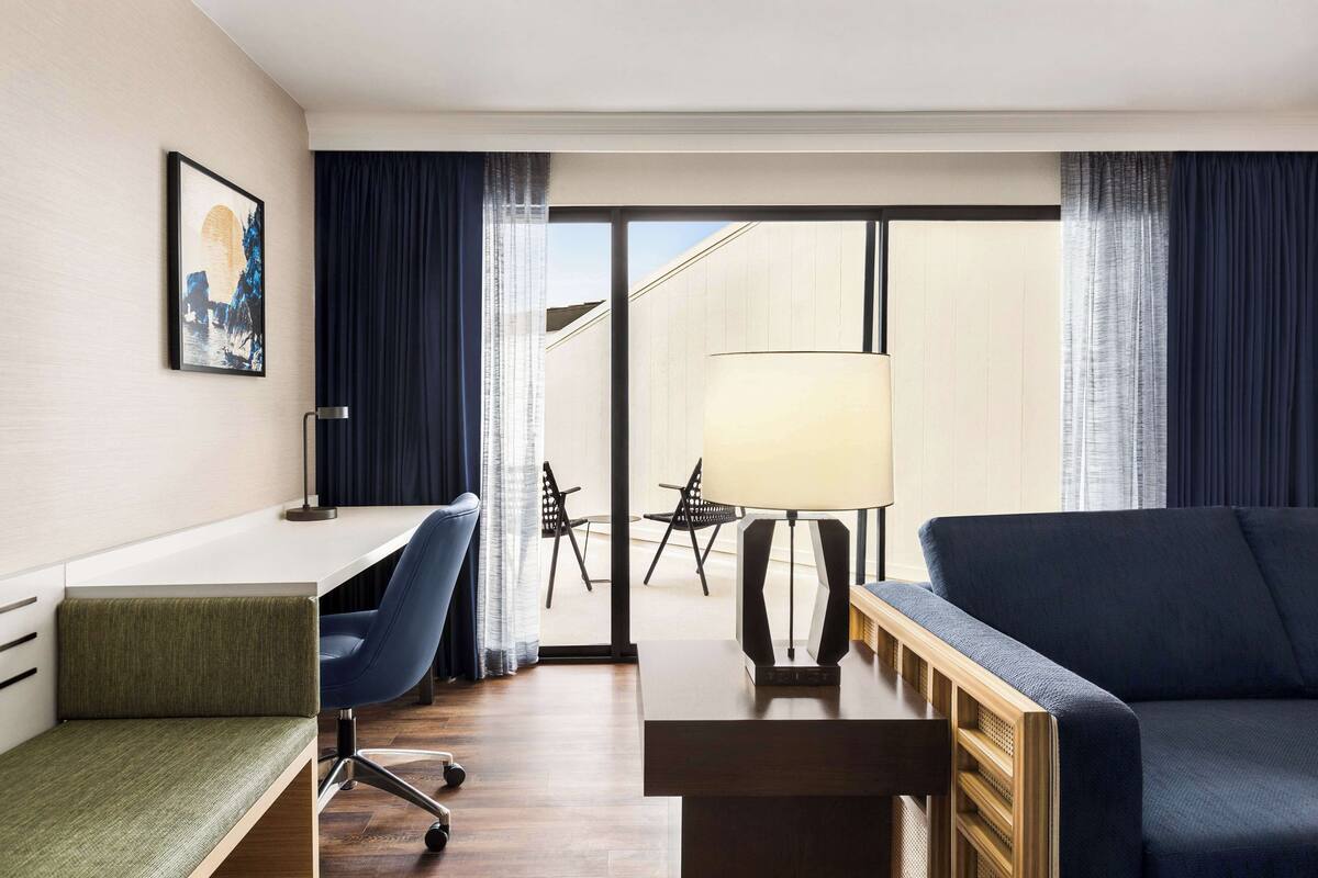 A hotel room with a desk, a blue chair, a sofa, a lamp, and a view of the outside.