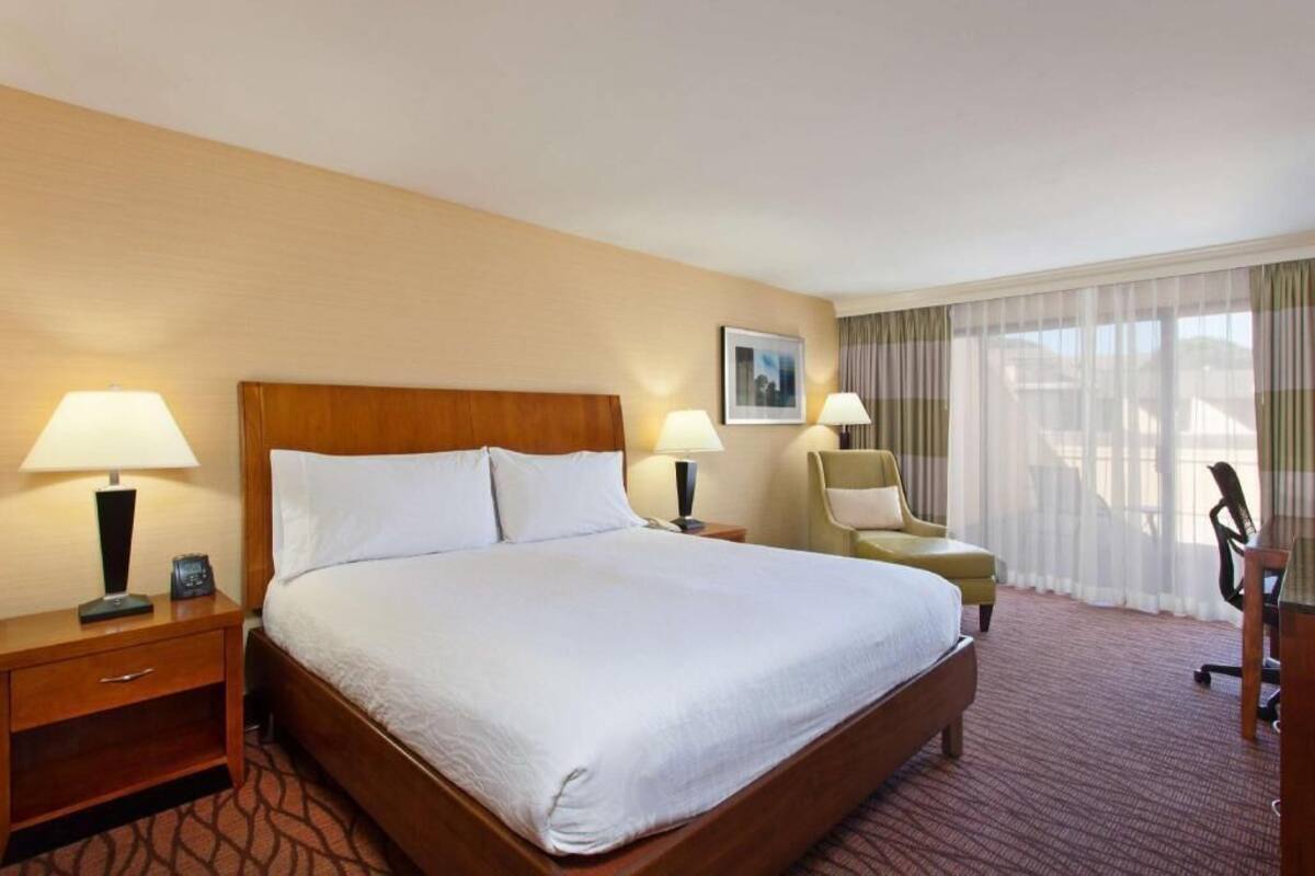 A hotel room with a large bed, two bedside tables with lamps, a chair, a desk, and a window with sheer curtains.