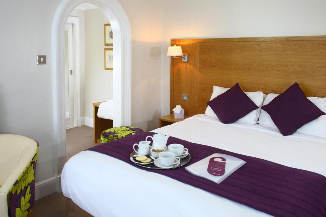 Habitación de hotel con una cama bien hecha, una bandeja con alimentos para el desayuno y una mesita de noche con lámpara.