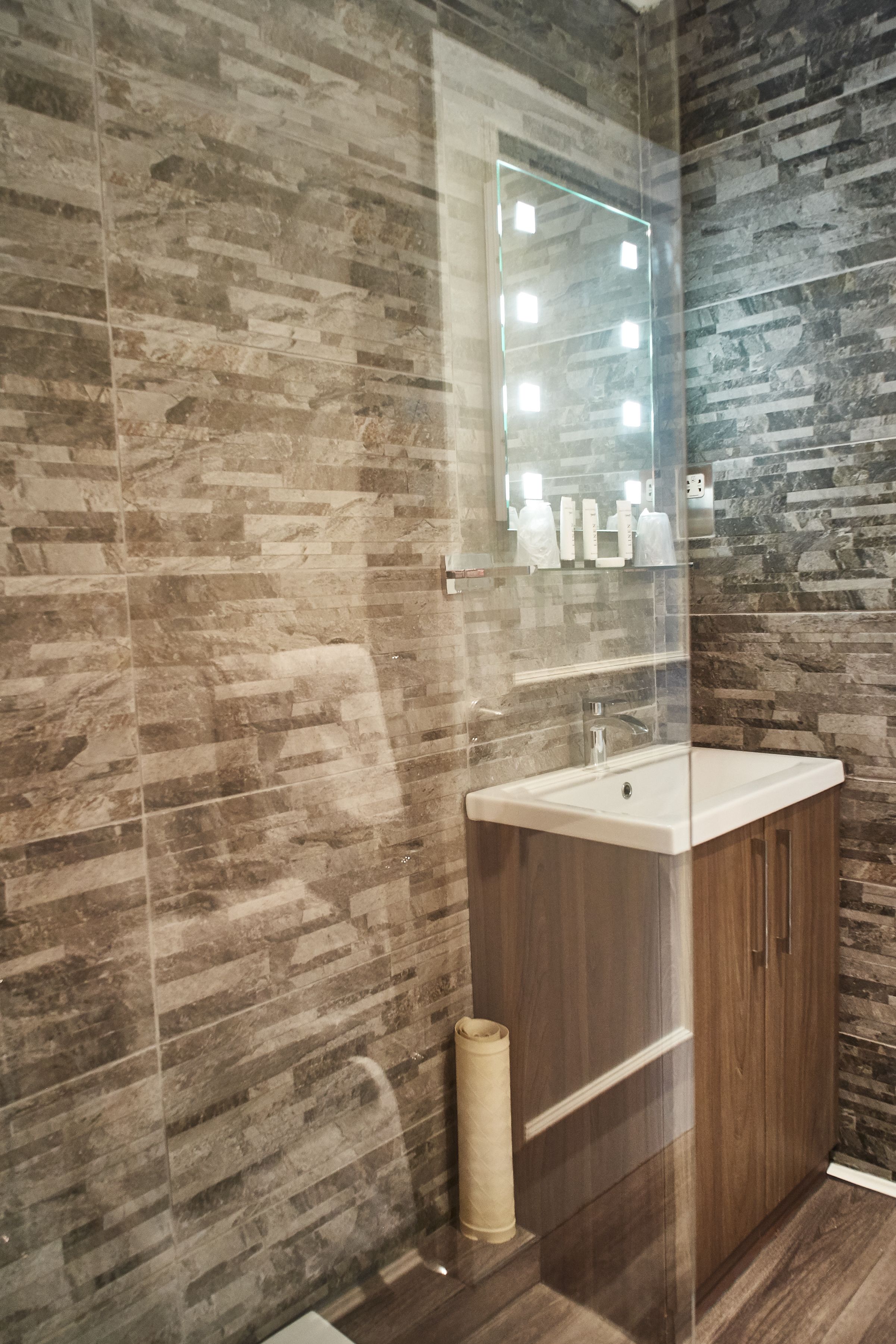 A bathroom with a stone-patterned wall, a glass-enclosed vanity, and a mirror with lights.