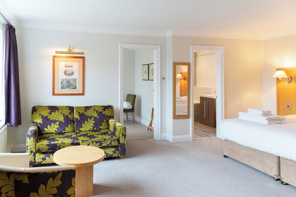A hotel room with a sofa, a bed, a small table, and a chair. There are framed pictures on the wall and a lamp on the wall.
