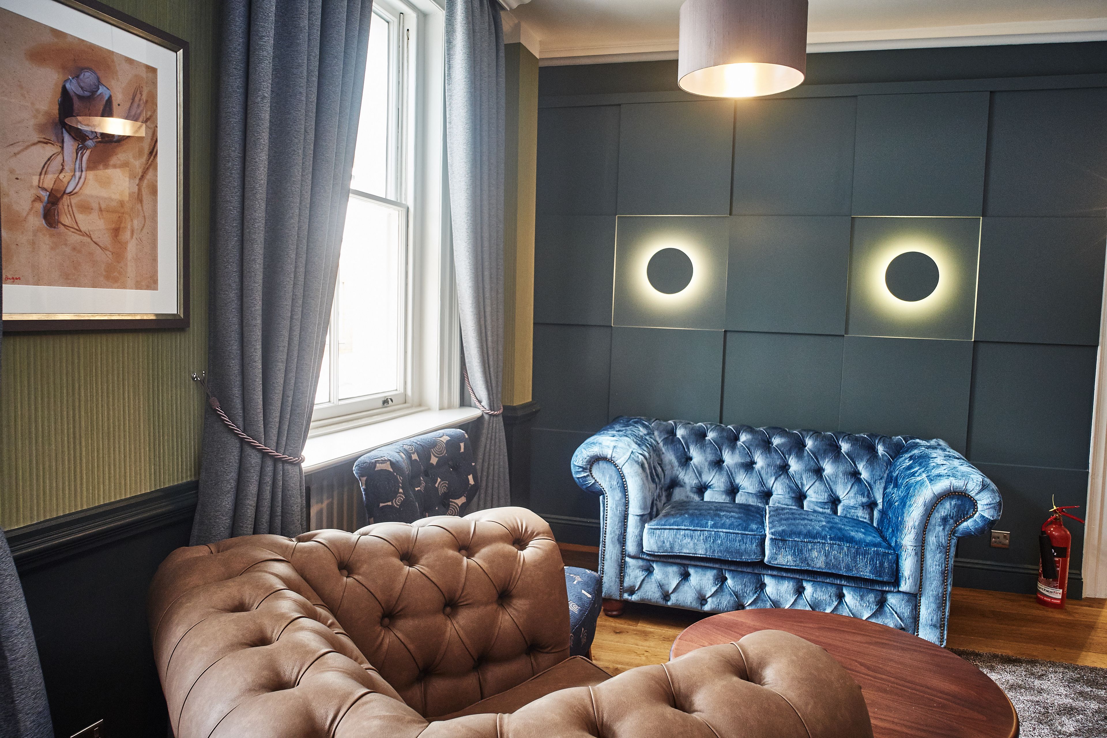 Een kamer met een bruine sofa met knoopjes, een blauwe sofa met knoopjes en een ronde houten salontafel. Er hangt een kunstwerk op de muur, een raam met gordijnen en een rond wandlamp.