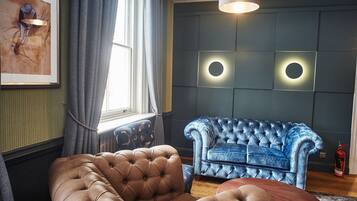 Een kamer met een bruine sofa met knoopjes, een blauwe sofa met knoopjes en een ronde houten salontafel. Er hangt een kunstwerk op de muur, een raam met gordijnen en een rond wandlamp.