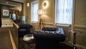 A modern living room with a glass partition, two black leather sofas, a round metallic ottoman, a floor lamp, and a radiator. - The Chequers Hotel (Newbury)