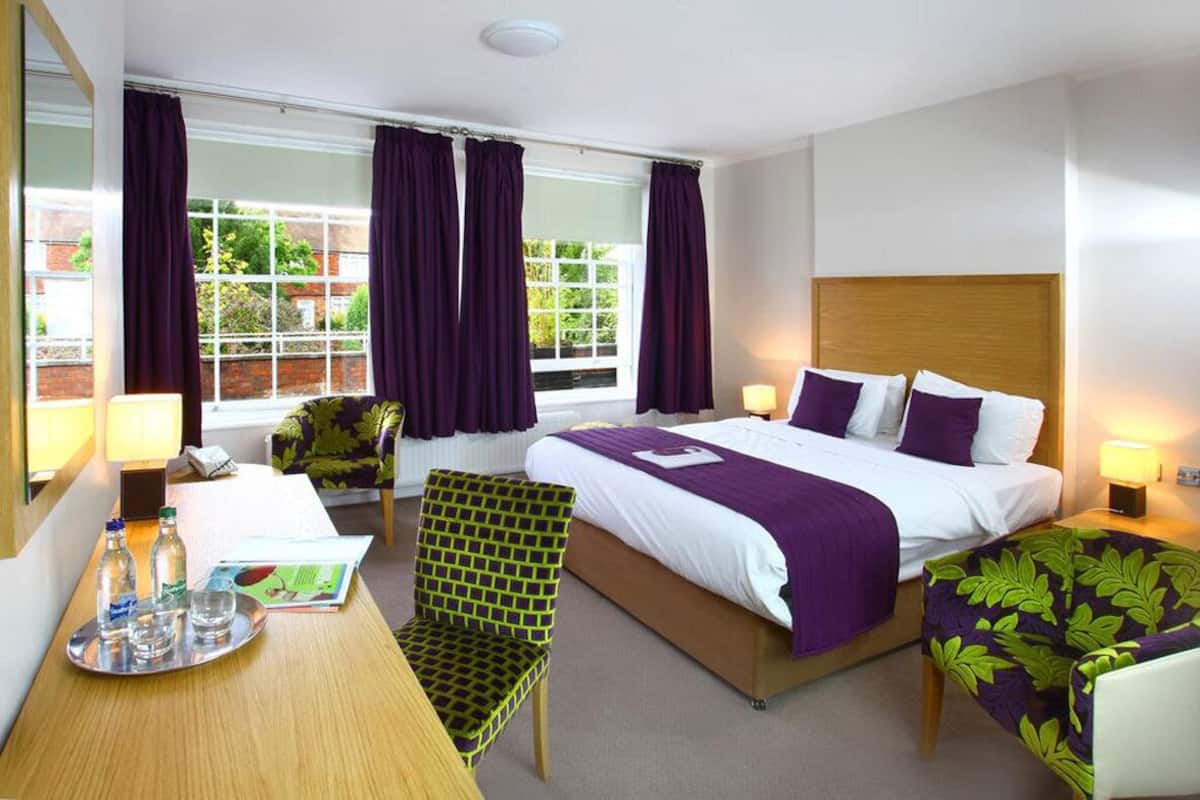 A hotel room with a large bed, a desk with a chair, a small table with a tray of items, and a window with curtains.