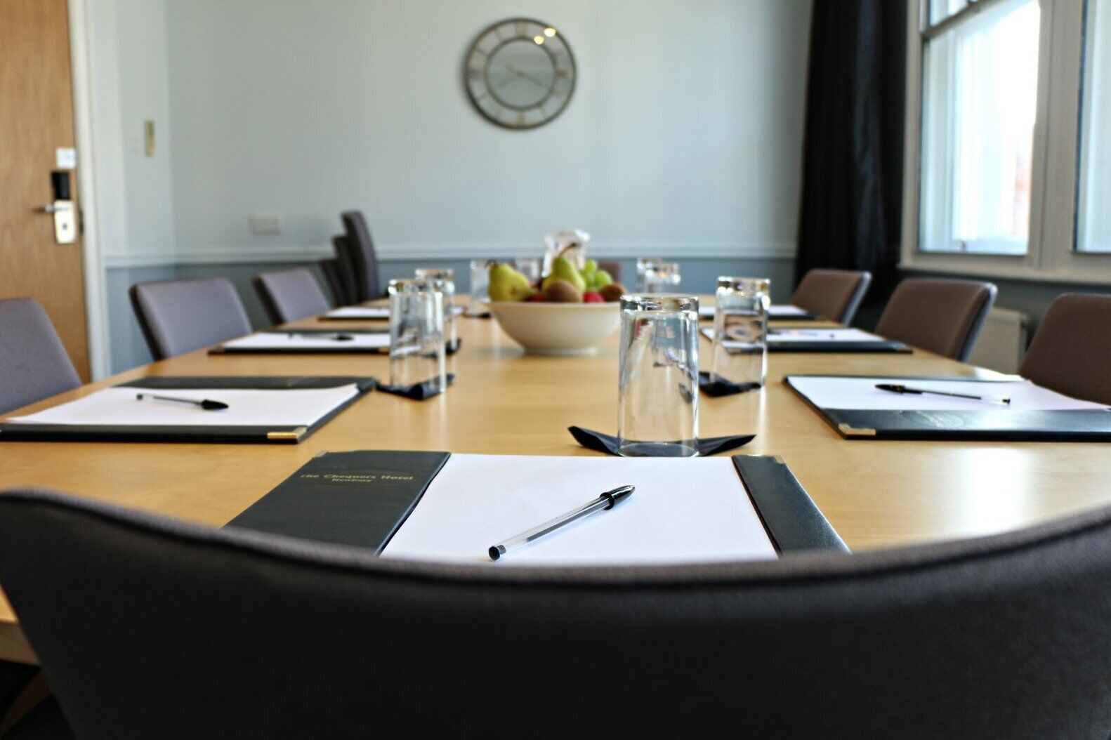 A conference room with a long table, chairs, a clock on the wall, and a bowl of fruit.