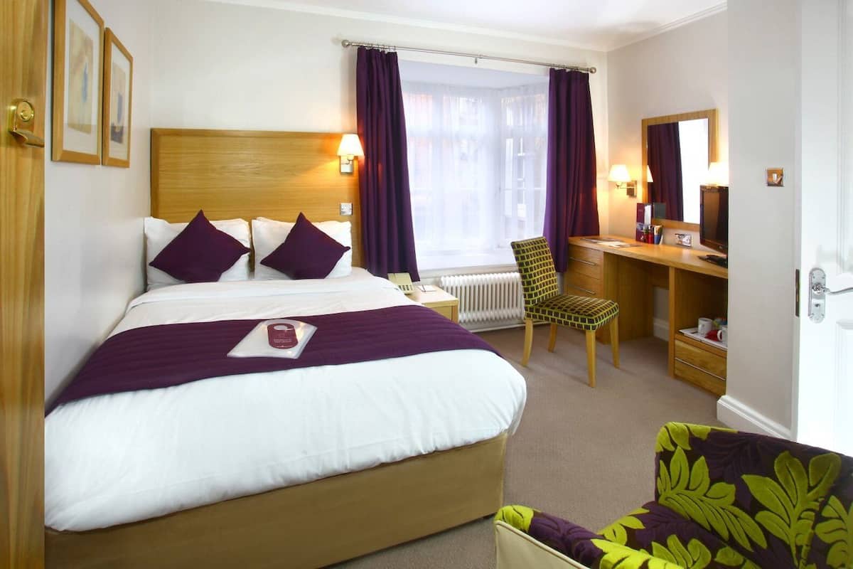 A hotel room with a large bed, a desk, a chair, and a window with curtains.