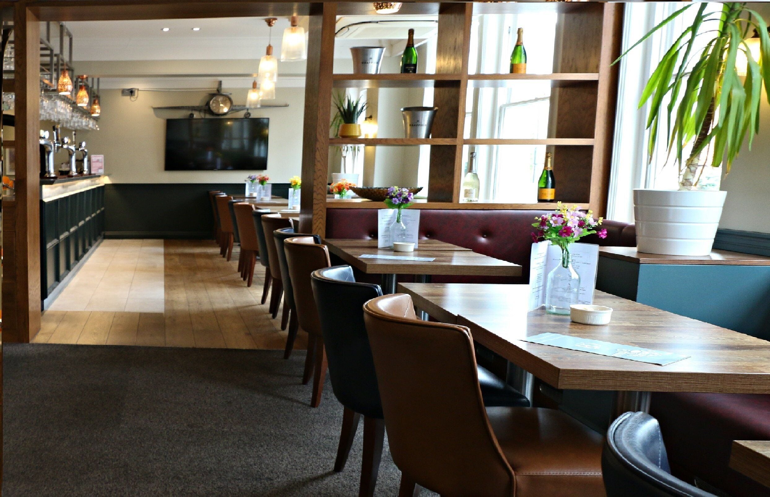 A restaurant with wooden tables and booths, a bar area with bottles, and a large potted plant.