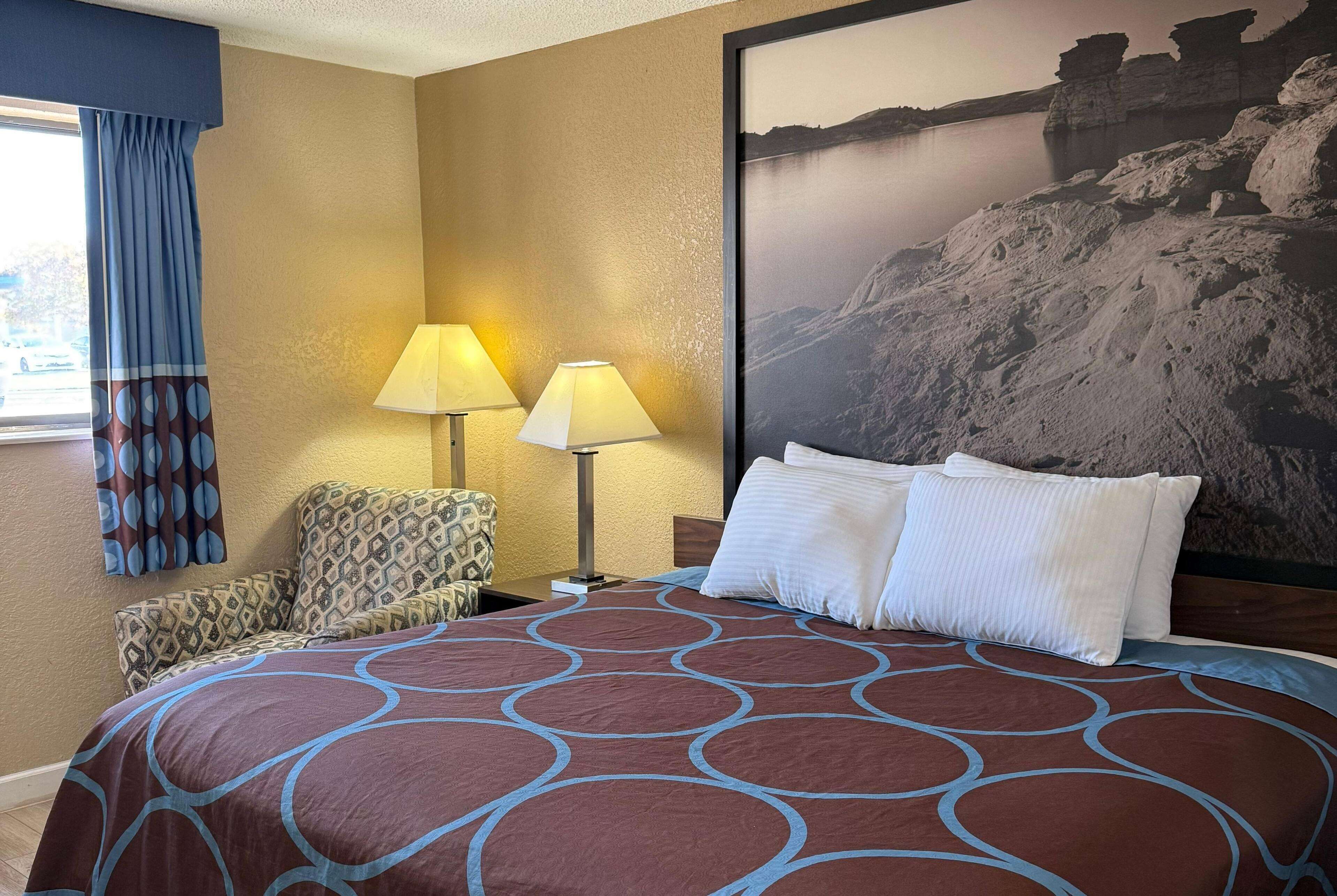 Habitación de hotel con una cama grande, dos lámparas, un sillón estampado y un cuadro grande en la pared.