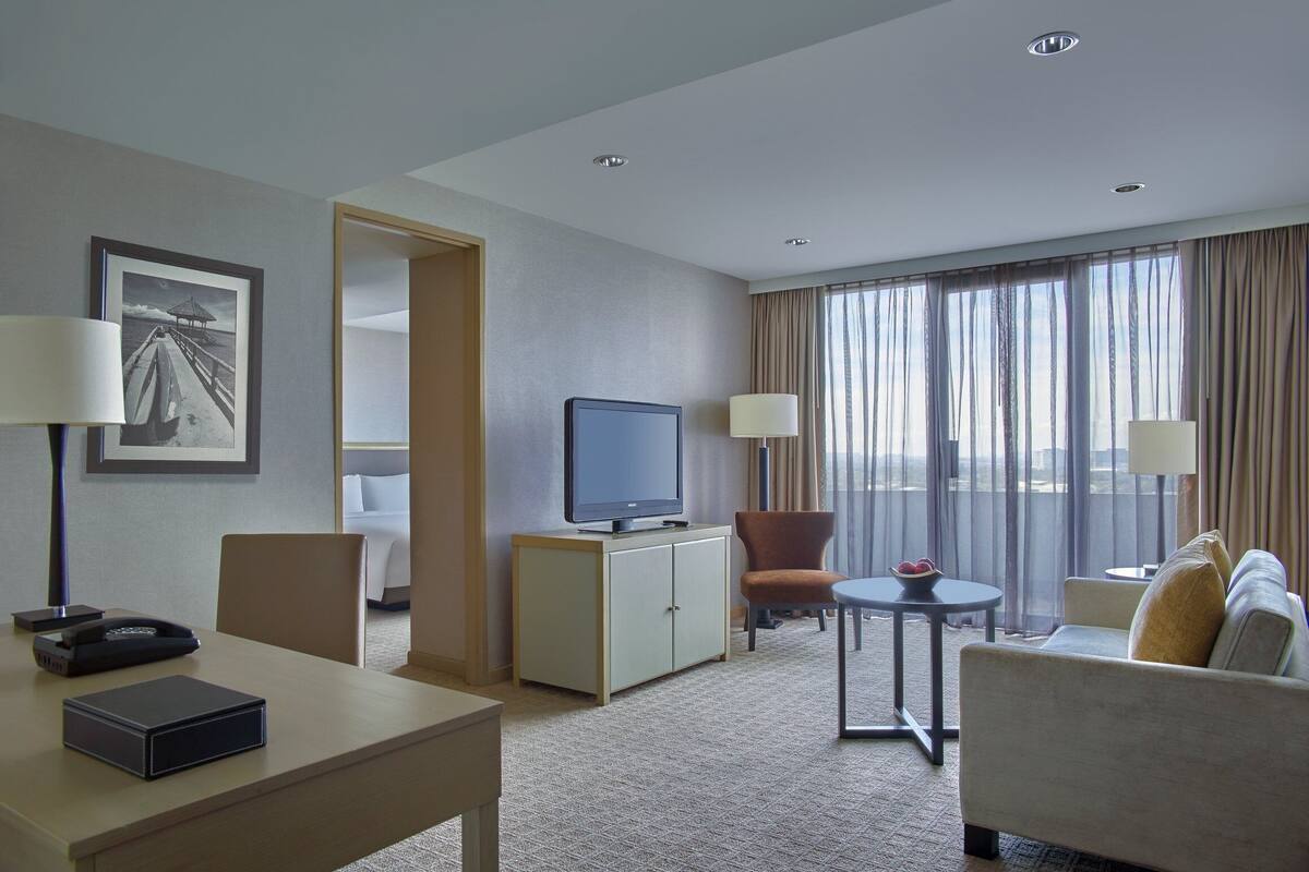Habitación de hotel con escritorio, silla, sofá, televisor y vistas a la ciudad a través de amplias ventanas.