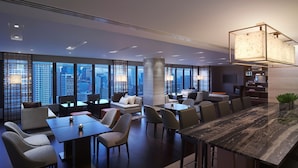 A modern hotel lobby with a high ceiling, large windows offering city views, a central seating area with sofas and tables, and a bar area with a unique geometric light fixture. - New World Makati Hotel (Makati)