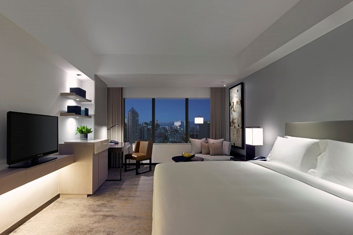 A modern hotel room with a large bed, a desk with a flat-screen TV, a seating area with a chair, and a city view through large windows.
