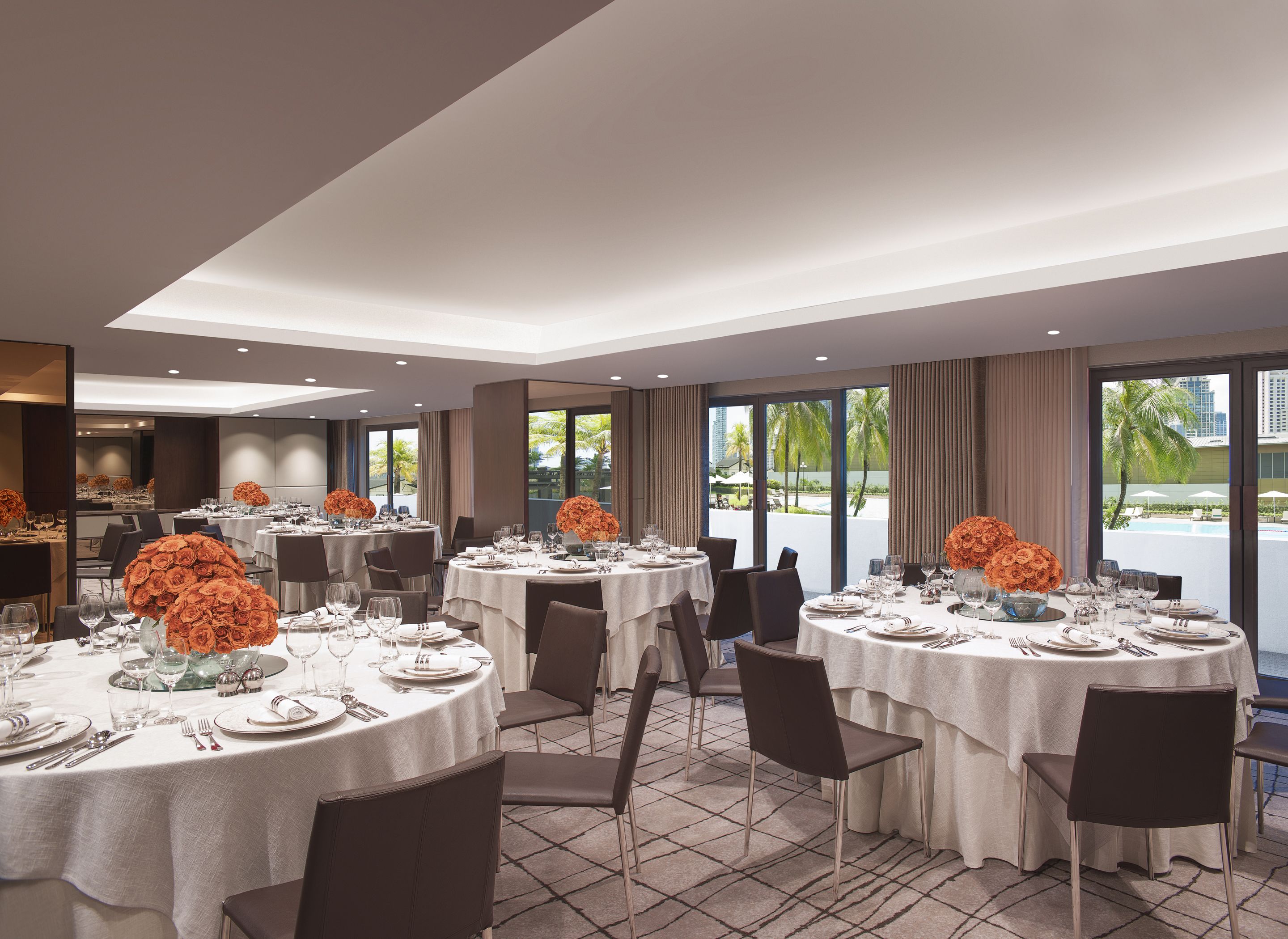 A hotel dining area with round tables set for service, large windows with a view of palm trees and a pool, and a modern interior design.