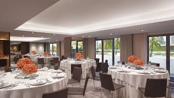 Un comedor de hotel con mesas redondas listas para el servicio, amplias ventanas con vistas a palmeras y una piscina, y un diseño interior moderno.