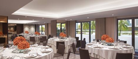 A hotel dining area with round tables set for service, large windows with a view of palm trees and a pool, and a modern interior design.