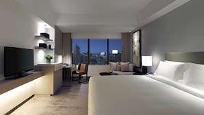 A modern hotel room with a large bed, a desk with a flat-screen TV, a chair, a small table with a plant, and a city view through large windows. - New World Makati Hotel (Makati)