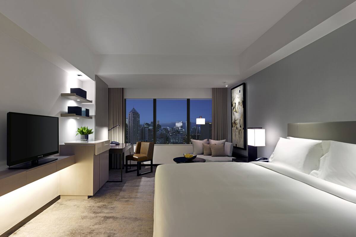 Una habitación de hotel moderna con una cama grande, un escritorio con televisor de pantalla plana, una silla, una mesita con una planta y vistas a la ciudad a través de amplias ventanas.