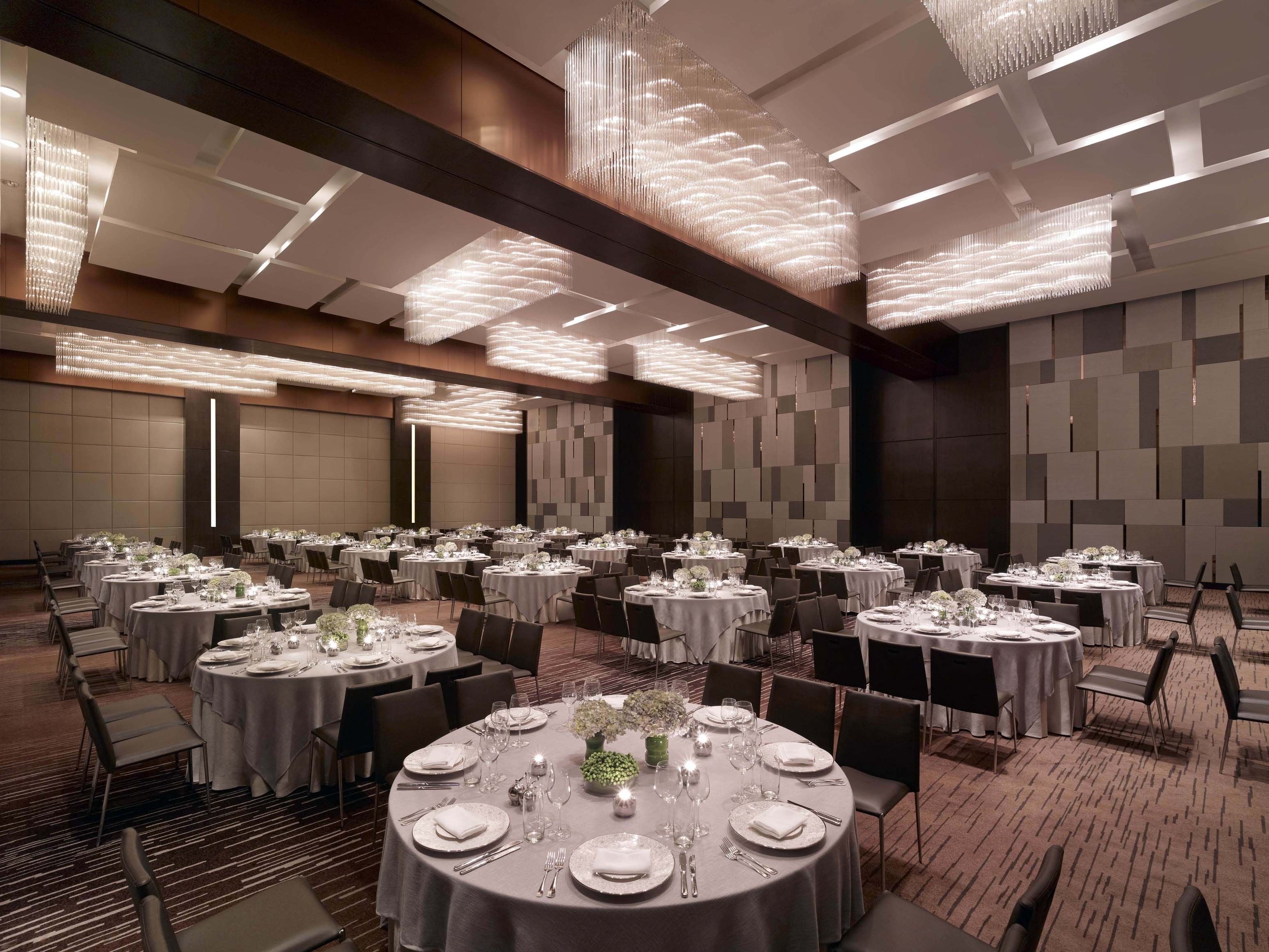 A spacious banquet hall with round tables set for dining, featuring modern chandeliers and a neutral color palette.