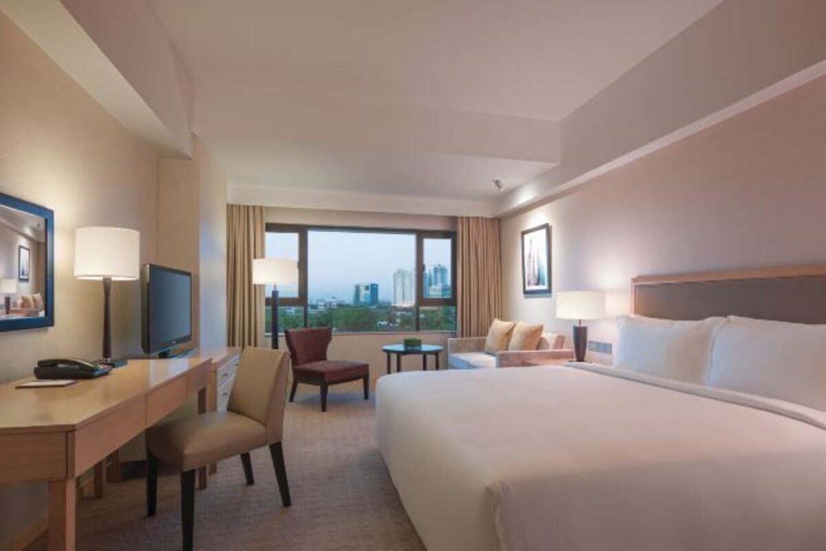 A hotel room with a large bed, a desk with a chair, a television, and a view of the cityscape through large windows.
