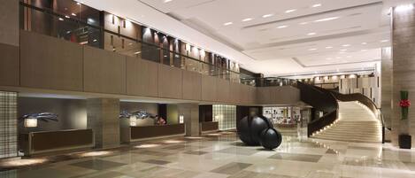 A spacious lobby with a high ceiling, a central staircase, and a reception area with modern furnishings.