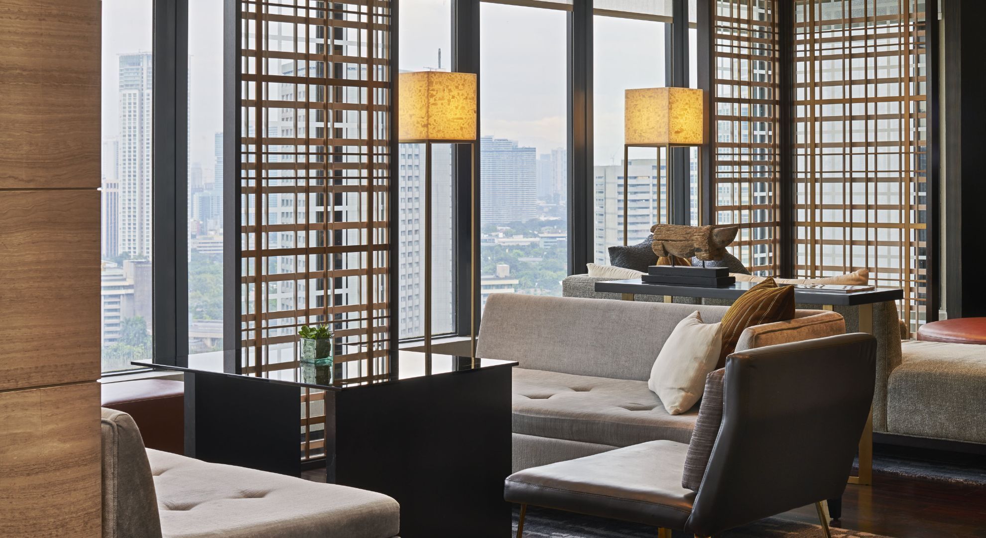 Habitación de hotel moderna con vistas a la ciudad, un sofá, sillas, una mesita con lámpara y objetos decorativos.